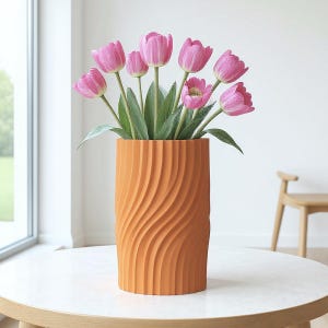 May include: A vibrant orange vase with a textured, swirling pattern, filled with pink tulips and green foliage. The vase is on a round table. The vase's height is approximately 25 cm.