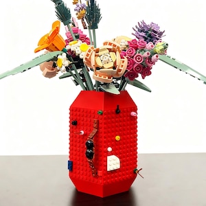 Può includere: Un vivace bouquet di fiori fatti con mattoncini di plastica colorati, disposti in un vaso di mattoni rossi. I fiori includono fiori arancioni, rosa e beige con foglie verdi. Il vaso ha una forma geometrica con borchie colorate.