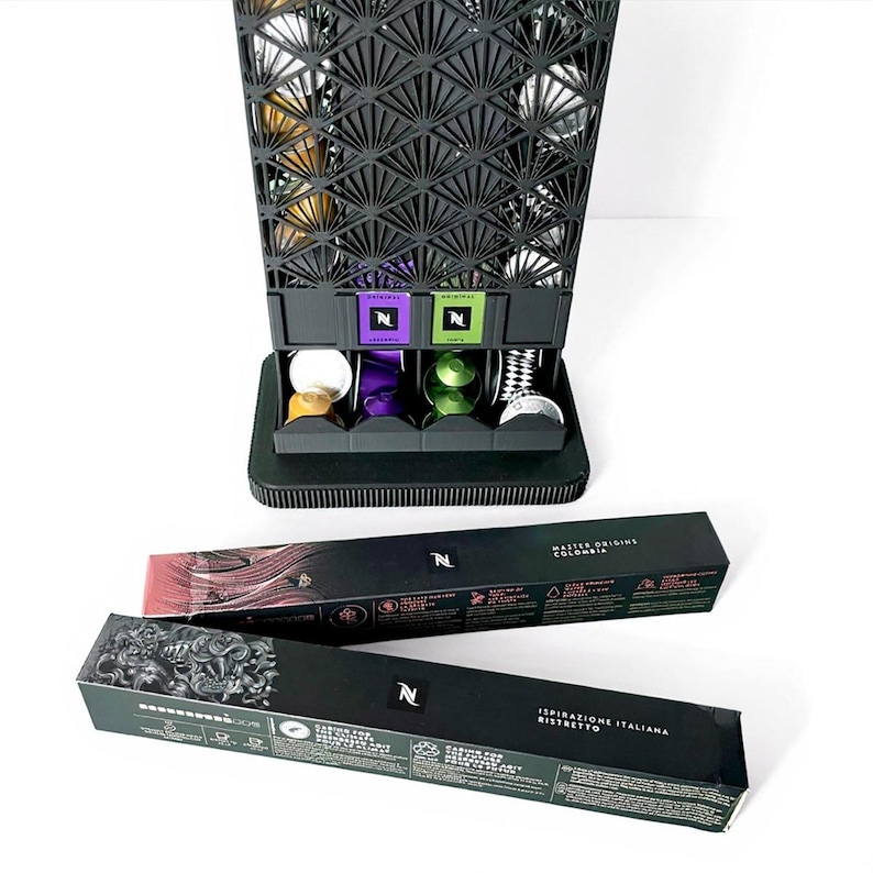 May include: A black coffee pod holder with a geometric design, filled with various coffee pods. Below, there are four pods in different colors. Two black boxes with text and the Nespresso logo are in the foreground.