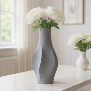 May include: A gray, modern vase with a unique, curved design, holding white peonies with green leaves. The vase sits on a white surface, with a smaller white vase of flowers in the background. A framed picture hangs on the wall.