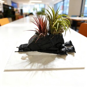 Può includere: Una fioriera nera stampata in 3D a forma di AT-AT Walker, riempita di vivaci piante aeree. La fioriera presenta dettagli meccanici ed è posizionata su una superficie bianca. Le piante aeree includono fogliame rosso e verde.