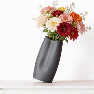 Könnte beinhalten: Eine graue Vase mit einem einzigartigen, geschwungenen Design hält einen lebendigen Strauß bunter Blumen. Die Blumen umfassen rote, rosa, orange, gelbe und weiße Blüten, die einen auffälligen Kontrast zum neutralen Farbton der Vase bilden. Die Vase steht auf einer weißen Oberfläche.