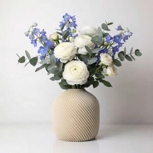 May include: A beige vase holding a floral display. The bouquet includes white ranunculus, blue delphiniums, and eucalyptus. The vase has a textured, ridged pattern. The flowers are arranged to create a full, lush display, suitable for home decoration.