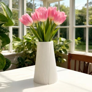 Könnte beinhalten: Eine weiße, plissierte Vase mit einem Strauß rosa Tulpen. Die Tulpen haben grüne Stiele und Blätter. Die Vase steht auf einem weißen Tisch vor einem Fenster mit Grünzeug im Freien.