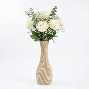 Könnte beinhalten: Eine beige Vase mit vertikalen Linien hält einen Strauß weißer Rosen, Schleierkraut und grüne Eukalyptusblätter. Die Vase hat eine schlanke, längliche Form, und die Blumen sind in voller Blüte. Die Anordnung ist vor weißem Hintergrund.