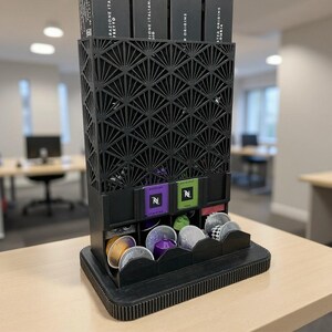 May include: Black coffee pod holder with a geometric design. The holder has multiple compartments for storing coffee pods. The top section holds boxes of coffee pods. The holder is made of a dark material and sits on a light-colored surface.
