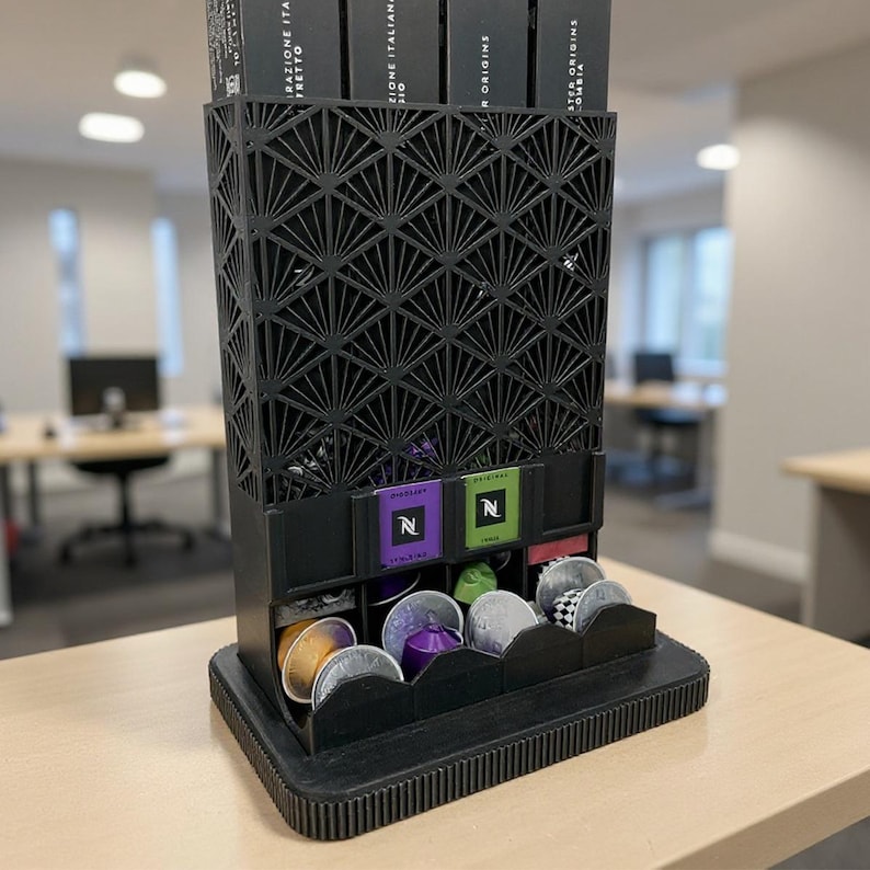 May include: Black coffee pod holder with a geometric design. The holder has multiple compartments for storing coffee pods. The top section holds boxes of coffee pods. The holder is made of a dark material and sits on a light-colored surface.