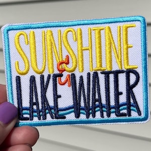 Puede incluir: Parche bordado con las palabras "SUNSHINE & LAKE WATER" en amarillo, azul y blanco. El parche tiene un borde azul y es de forma rectangular. El parche se sostiene sobre un fondo blanco.