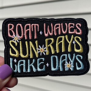 Könnte beinhalten: Gestickter Aufnäher mit den Worten "BOAT.WAVES SUN.RAYS LAKE.DAYS" in Rosa, Gelb und Blau. Der Aufnäher hat einen schwarzen Rand und kleine Blumenakzente. Der Aufnäher wird vor einem weißen Hintergrund gehalten.