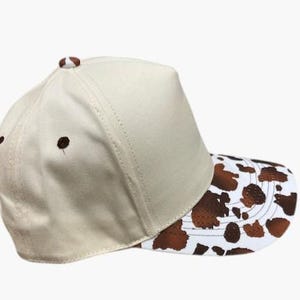 May include: A beige baseball cap with a brown and white cow print on the brim. The cap has a curved brim and a button on top. The cap is made of a sturdy material.