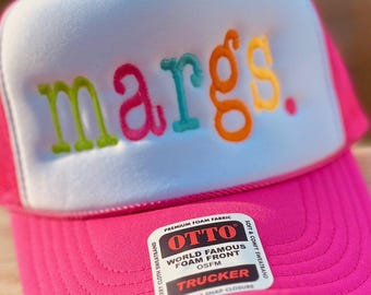 May include: A pink trucker hat with a white front panel. The front panel has the word "margs." embroidered in colorful letters. The hat has a black mesh back and a black snap closure.