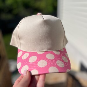 May include: A beige baseball cap with a pink brim featuring white polka dots. The cap is held up against a blurred green background. The cap's crown is a solid beige color.