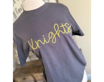 May include: A dark gray t-shirt with the word "Knights" embroidered in yellow thread.