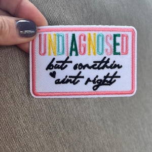 May include: Embroidered patch with the words "UNDIAGNOSED" in colorful letters, and the phrase "but somethin ain't right" in black script. The patch has a white background with a pink border.