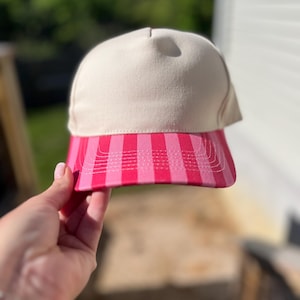 May include: A baseball cap with a cream-colored crown and a pink and red striped brim. The cap is held up in front of a blurred green and white background.