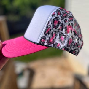 May include: A trucker hat with a white front panel, a pink brim, and a mesh leopard print design on the back. The hat has a black rope detail and is angled to the right.