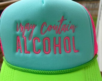 May include: A neon green and turquoise trucker hat with the embroidered text "may contain ALCOHOL" in pink. The hat has a pink mesh back and a green braided trim.
