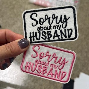 Puede incluir: Dos parches rectangulares bordados con el texto "Sorry about my HUSBAND". Uno tiene fondo blanco con texto y borde negros. El otro tiene fondo blanco con texto y borde rosas. Los parches están sujetos por una mano.