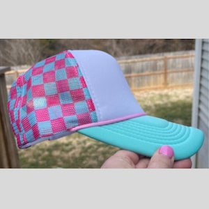 May include: A trucker hat with a light blue brim and a white front panel. The mesh back features a pink and light blue checkered pattern. A pink cord accents the brim.