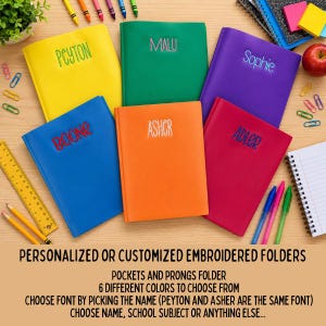 May include: Six colorful folders in yellow, green, purple, blue, orange, and pink, each featuring a name embroidered on the front cover. The folders are arranged on a surface alongside a yellow ruler, colored pencils, and a spiral notebook.