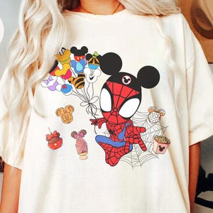 Puede incluir: Camiseta de color crema con un personaje de dibujos animados de Spider-Man con orejas de Mickey Mouse, sosteniendo globos con caras de personajes de Disney. El dise&ntilde;o incluye ilustraciones de comida y una telara&ntilde;a. La palabra "lovry" est&aacute; en un c&iacute;rculo en la parte superior izquierda.