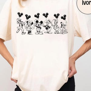 May include: Ivory-colored t-shirt featuring a black and white illustration of Disney characters holding Mickey Mouse-shaped balloons. The characters include Goofy, Minnie Mouse, Mickey Mouse, Donald Duck, Daisy Duck, and Pluto. The word "Ivory" is in a circle.