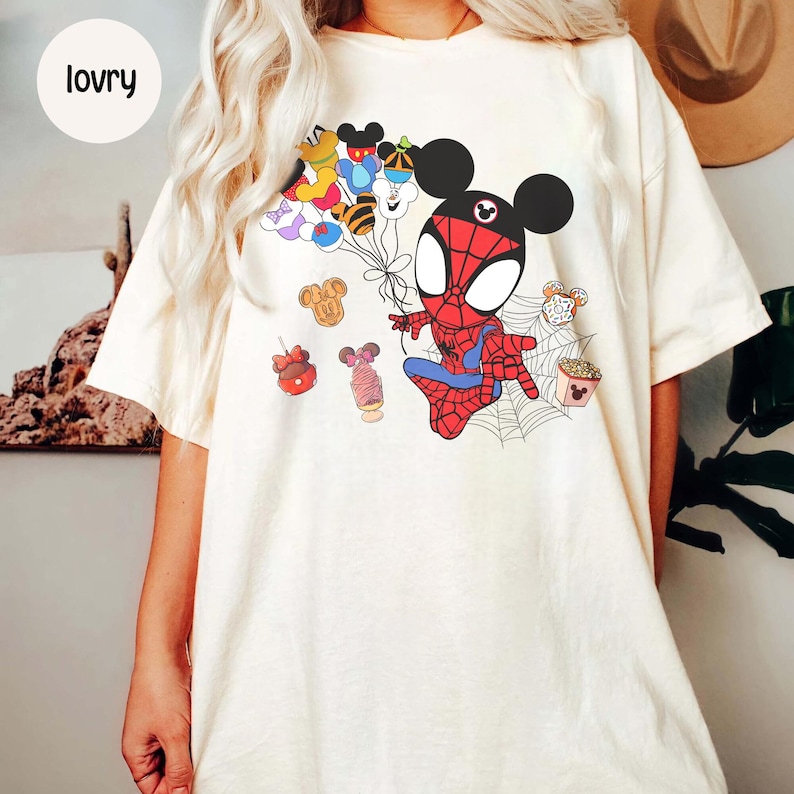 Puede incluir: Camiseta de color crema con un personaje de dibujos animados de Spider-Man con orejas de Mickey Mouse, sosteniendo globos con caras de personajes de Disney. El dise&ntilde;o incluye ilustraciones de comida y una telara&ntilde;a. La palabra "lovry" est&aacute; en un c&iacute;rculo en la parte superior izquierda.