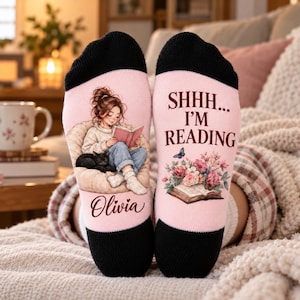 May include: A pair of light pink socks with black cuffs. The left sock features an illustration of a person reading a book with a black cat, and the name Olivia. The right sock displays the text SHHH... I'M READING above a floral arrangement and an open book.