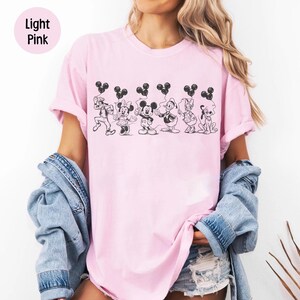 May include: Light pink t-shirt featuring a black and white graphic of Disney characters holding Mickey Mouse-shaped balloons. The characters include Mickey Mouse, Minnie Mouse, Donald Duck, Daisy Duck, Goofy, and Pluto. The words "Light Pink" are in a pink circle in the upper left corner.