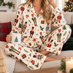 May include: A two-piece pyjama set featuring a long-sleeved button-down shirt and matching trousers in an off-white fabric. The garment is decorated with a repeating pattern of nutcrackers, Christmas trees, gift boxes, and holly leaves.