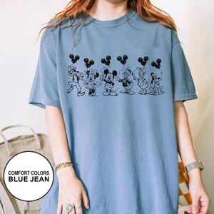 May include: A blue jean Comfort Colors t-shirt featuring a black line drawing of Disney characters holding Mickey Mouse-shaped balloons. The characters include Goofy, Minnie Mouse, Mickey Mouse, Donald Duck, Daisy Duck, and Pluto.