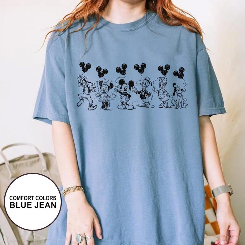 May include: A blue jean Comfort Colors t-shirt featuring a black line drawing of Disney characters holding Mickey Mouse-shaped balloons. The characters include Goofy, Minnie Mouse, Mickey Mouse, Donald Duck, Daisy Duck, and Pluto.