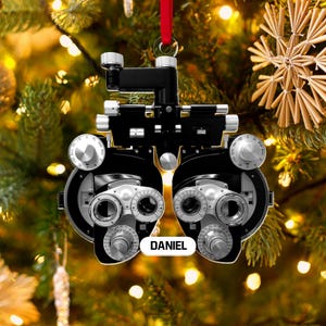 May include: A black and silver phoropter ornament with the name "DANIEL" on a white oval. The ornament hangs from a red ribbon against a backdrop of a Christmas tree with lights and a star-shaped decoration.