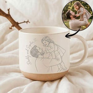 May include: Cream-colored ceramic mug with a brown base and a large handle. The mug features a sketch of a father and child, with the words "Daddy and Sophia" in script. A circular photo of the pair is also included.