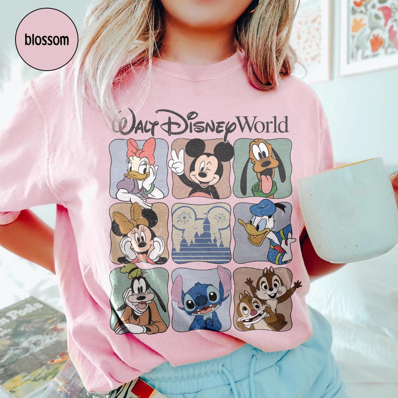 Pu&ograve; includere: T-shirt rosa chiaro con un motivo di personaggi Disney, tra cui Topolino, Minnie, Paperino e Pippo. La scritta "Walt Disney World" &egrave; stampata sopra in stile vintage.