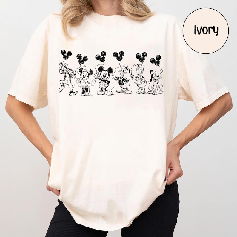 May include: Ivory-colored t-shirt featuring a black and white illustration of Disney characters holding Mickey Mouse-shaped balloons. The characters include Goofy, Minnie Mouse, Mickey Mouse, Donald Duck, Daisy Duck, and Pluto. The word "Ivory" is in a circle.