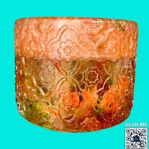 May include: A decorative, cylindrical container with an intricate floral pattern. The resin-based piece features a gradient of orange and gold hues, with embedded glitter and floral accents. The bottom half has a detailed embossed design. The words "SCAN ME" and a QR code are visible.