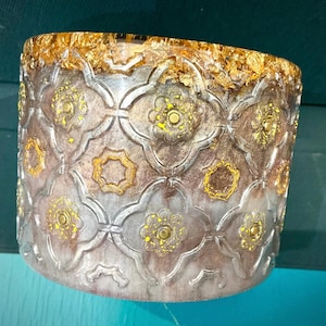 May include: A decorative cylindrical container with a gold leaf rim and a patterned design. The design features repeating floral and geometric shapes in gold and silver tones. The container has a textured surface.