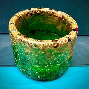 May include: A handcrafted, cylindrical container in a vibrant green shade, featuring a decorative pattern. The rim is embellished with gold leaf and colourful accents. The container is likely used for holding small items or as a decorative piece.