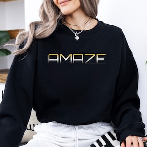 May include: Black crewneck sweatshirt with the word "AMAZE" printed across the chest in gold and silver lettering. The sweatshirt is a solid black color and has long sleeves. The overall style is casual and comfortable.