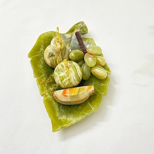 May include: A stone fruit arrangement featuring a pear, an apple, a bunch of grapes, and a fruit slice, presented on a green leaf-shaped base. The pear and apple have gold detailing. The arrangement is made of stone.