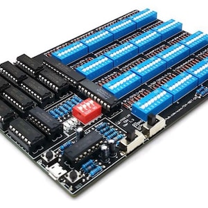May include: A black circuit board with various electronic components. It features rows of blue and black connectors, red switches, and resistors and chips. The board is designed for electronic projects.