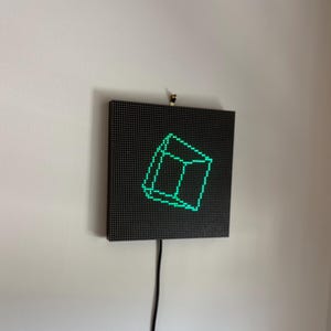 May include: A square black LED pixel display panel mounted on a wall. The screen features a bright green glowing wireframe cube graphic composed of individual light dots. A thin black power cable extends downward from the bottom of the device.