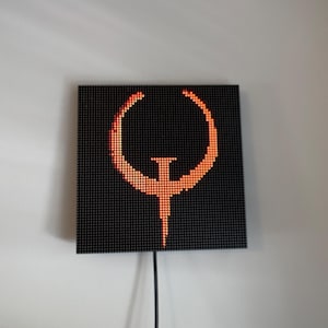 May include: A square LED matrix display panel featuring a glowing orange pixelated Quake logo centred against a black background. The device is mounted on a wall and includes a black power cable extending downwards from the bottom centre edge.