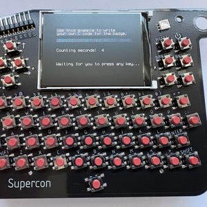 Pode incluir: Dispositivo eletrônico preto com uma pequena tela exibindo texto. Possui inúmeros botões vermelhos dispostos como um teclado, com a palavra "Supercon" impressa na parte inferior. A tela mostra instruções para escrever código.