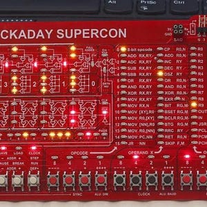 Puede incluir: Una placa de circuito rojo con el texto "HACKADAY SUPERCON" impreso en ella. La placa tiene una variedad de LED, interruptores y otros componentes. Los LED están encendidos en un patrón que deletrea la palabra "HACKADAY".