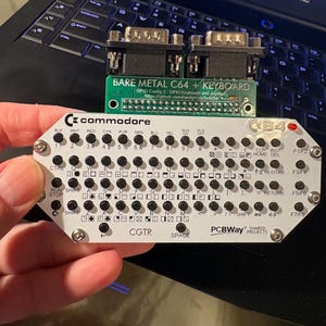 May include: A Commodore C64 keyboard with a green circuit board labeled "BARE METAL C64 + KEYBOARD" and a black laptop keyboard in the background. The C64 keyboard has various keys and labels, including "CTRL", "SHIFT", and "SPACE".