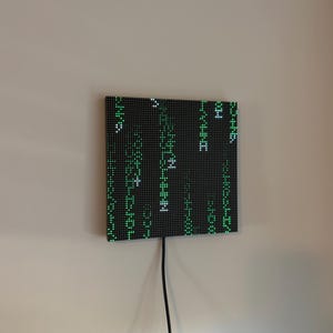 Puede incluir: Una pantalla LED cuadrada de pared con un panel de rejilla negro y caracteres digitales verdes y blancos iluminados dispuestos en columnas verticales, que recuerdan a código informático, con un cable de alimentación negro en la parte inferior.