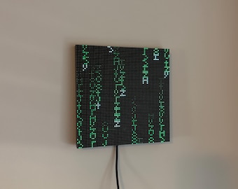 May include: A square LED wall display featuring a black grid panel with illuminated green and white digital characters arranged in vertical columns, resembling falling code, with a black power cord extending from the bottom center.