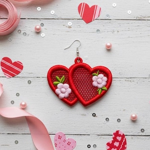 Pode incluir: Brincos vermelhos em forma de coração com bordado floral, exibidos com fita rosa, recortes em forma de coração e lantejoulas em uma superfície de madeira branca. Os brincos apresentam uma textura semelhante a uma malha.